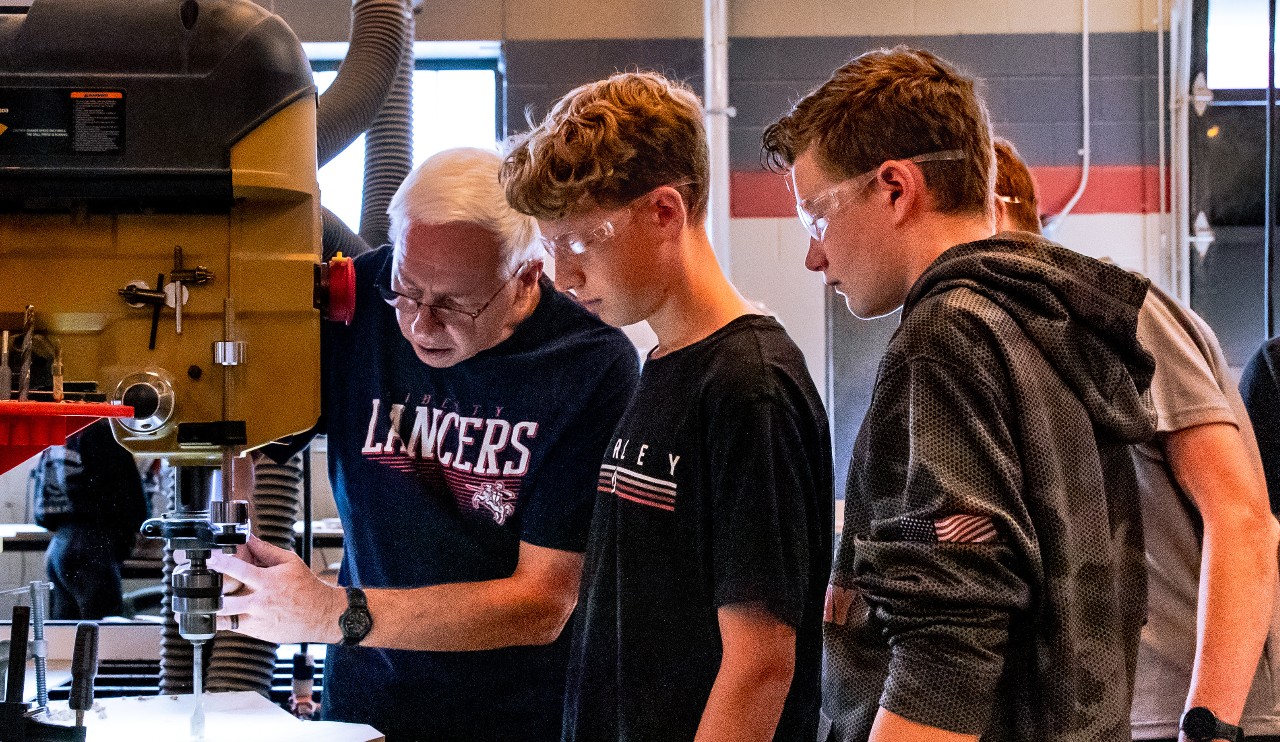 Two students watch as a teacher demonstrates the use of a drill press.